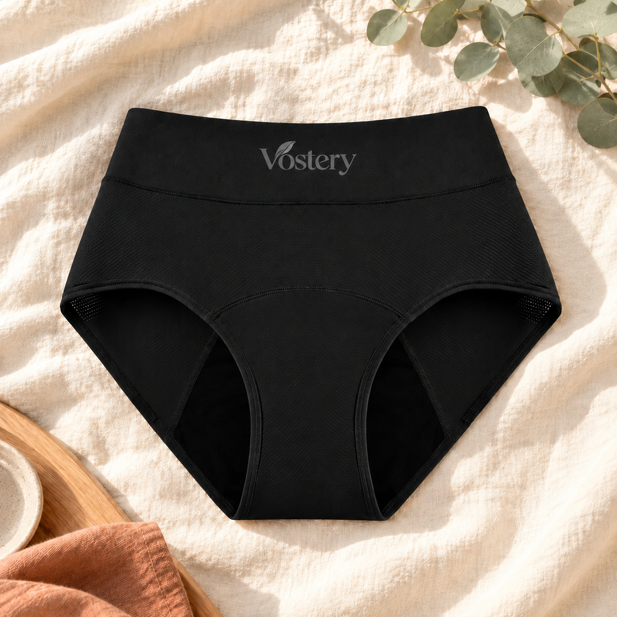 Culotte absorbante Vostery noire taille haute posée à plat sur du lin, avec panneaux latéraux en filet